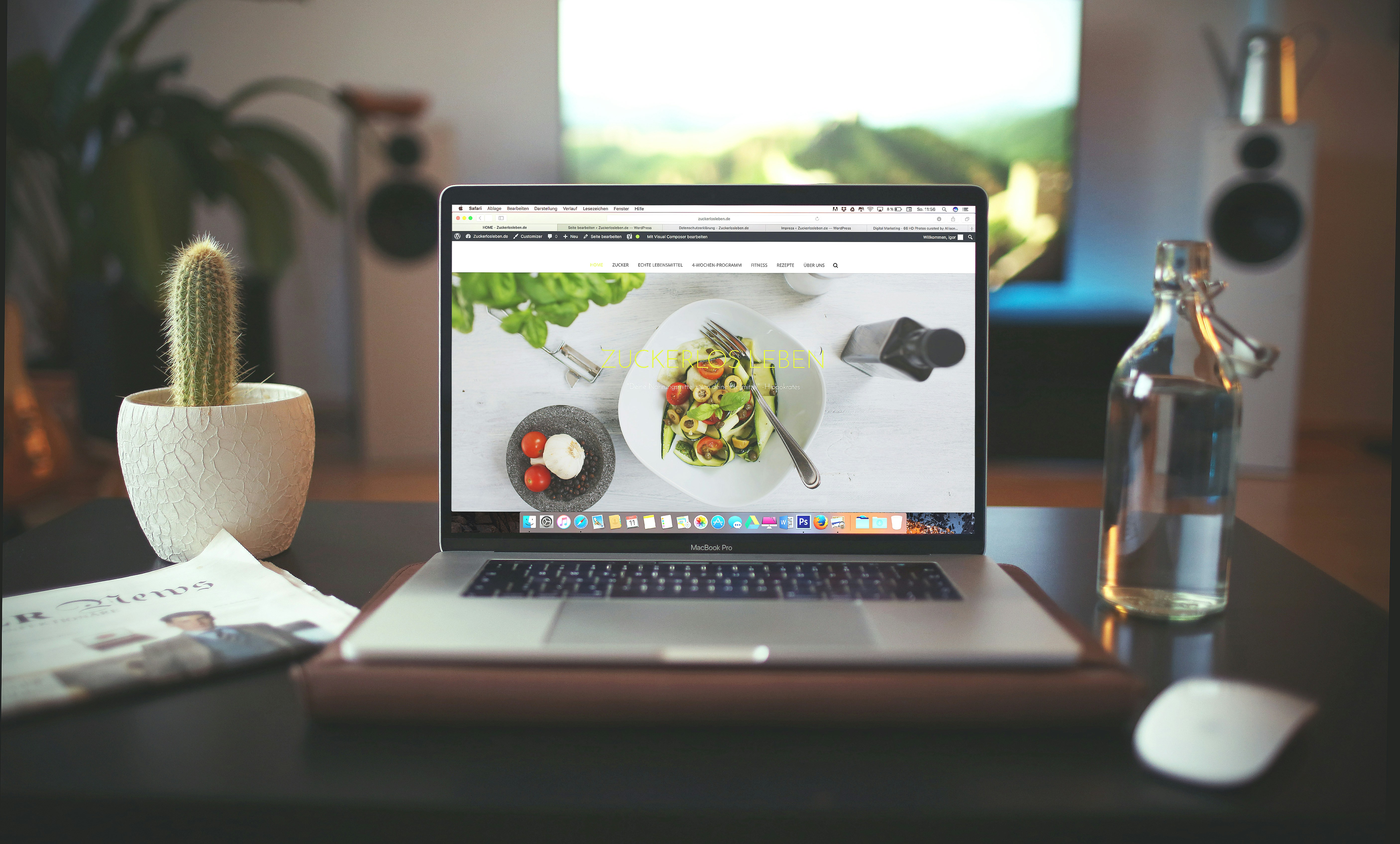 A laptop displaying a food-related webpage with a bowl of salad and ingredients in the foreground, alongside a small cactus, a glass bottle of water, and a magazine on a desk.